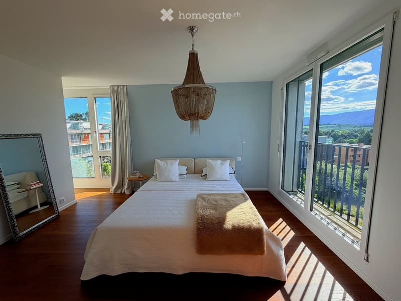 Bright bedroom with wooden flooring, large windows offering a scenic view, a neatly made bed with pillows and a blanket, a mirror, a small table, and a chandelier.