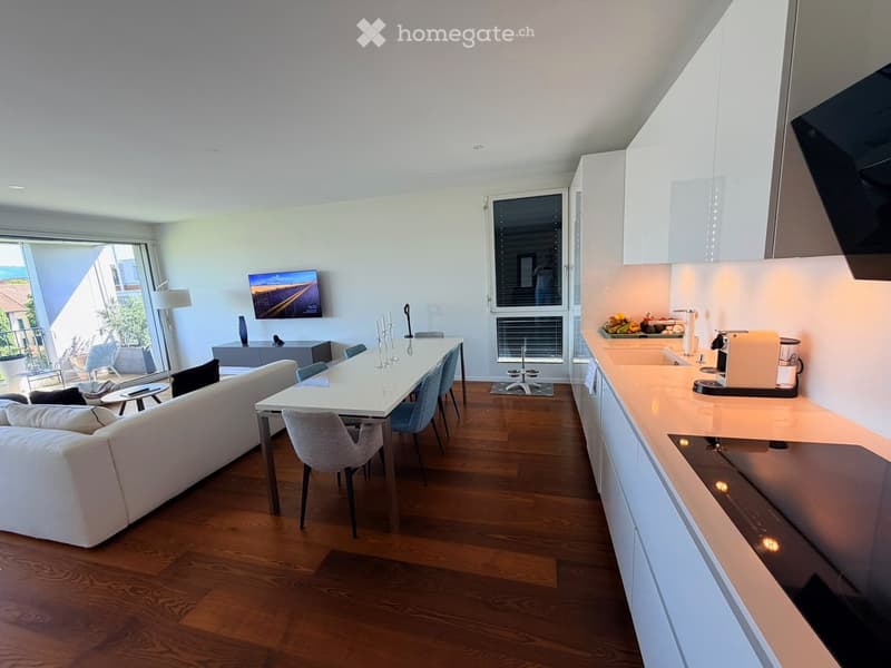 Open-plan living and dining area with a white sofa, dining table, chairs, and a modern kitchen counter with appliances. Large windows provide natural light, and a television is mounted on the wall.