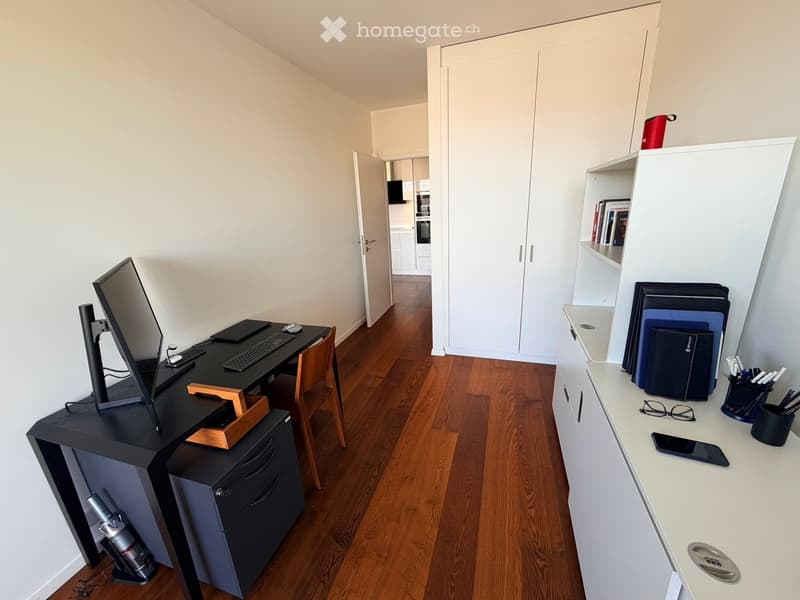 A modern, well-lit room with wooden flooring, a black desk with a screen, keyboard, and mouse, a white shelf with books and a laptop, and a small white cabinet.