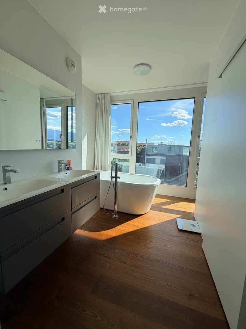 Modern bathroom with wooden flooring, a freestanding bathtub, double sink vanity, large windows offering a scenic view, and a mirror above the sink.