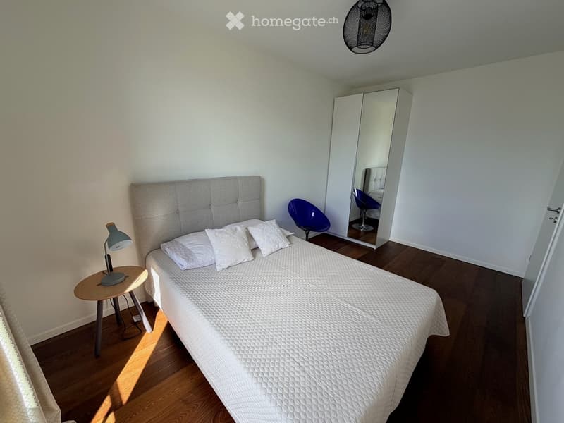 Modern bedroom with a white bed, grey headboard, white pillows, a nightstand with a lamp, a blue chair, and a large mirrored wardrobe.