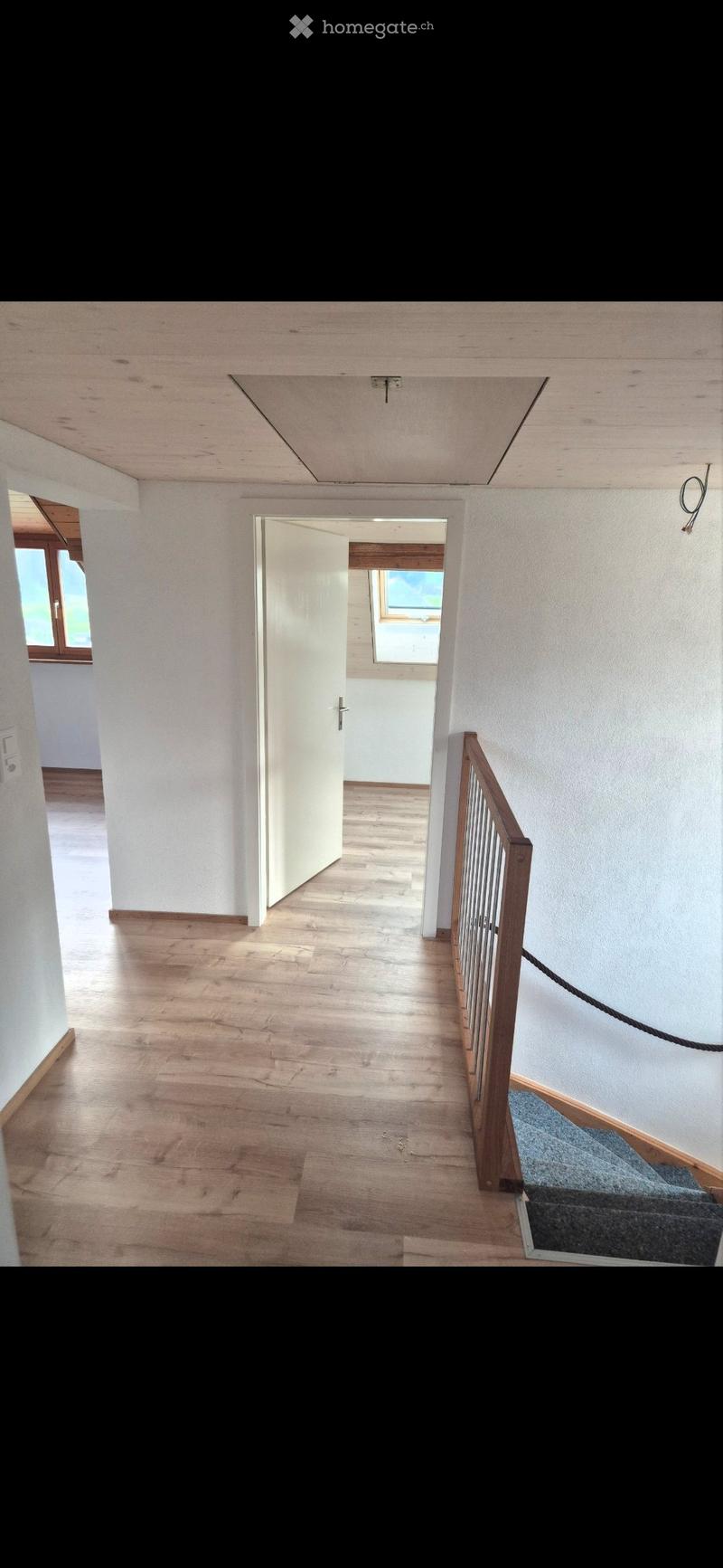 Empty hallway with wooden flooring, white walls, and a staircase leading upwards. A door is open to another room with a window visible.