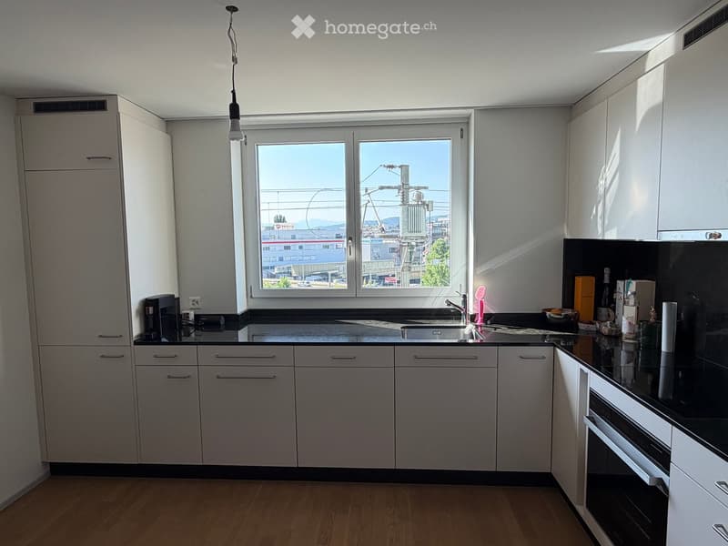 Modern kitchen with white cabinets, black countertops, and a large window overlooking the city. Includes a sink, faucet, oven, coffee maker, and various small items on the counter.