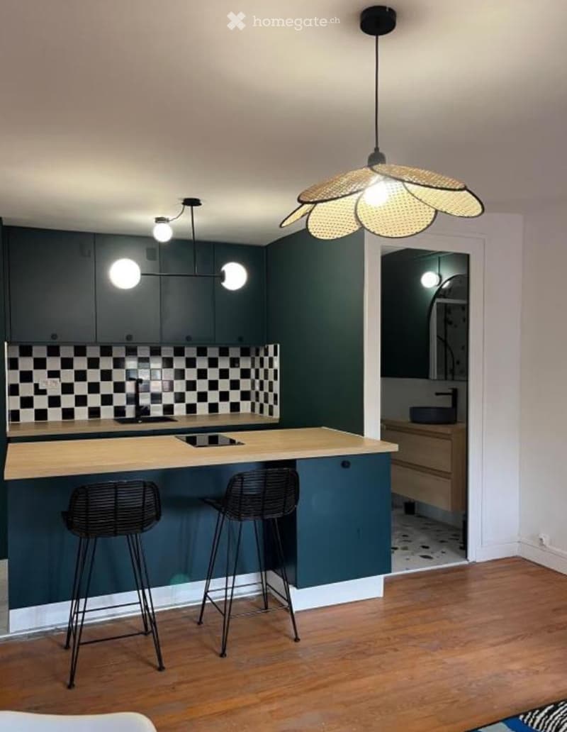 Modern kitchen with dark green cabinetry, a checkered backsplash, a wooden island, and two black bar stools. A large pendant light hangs above the island.