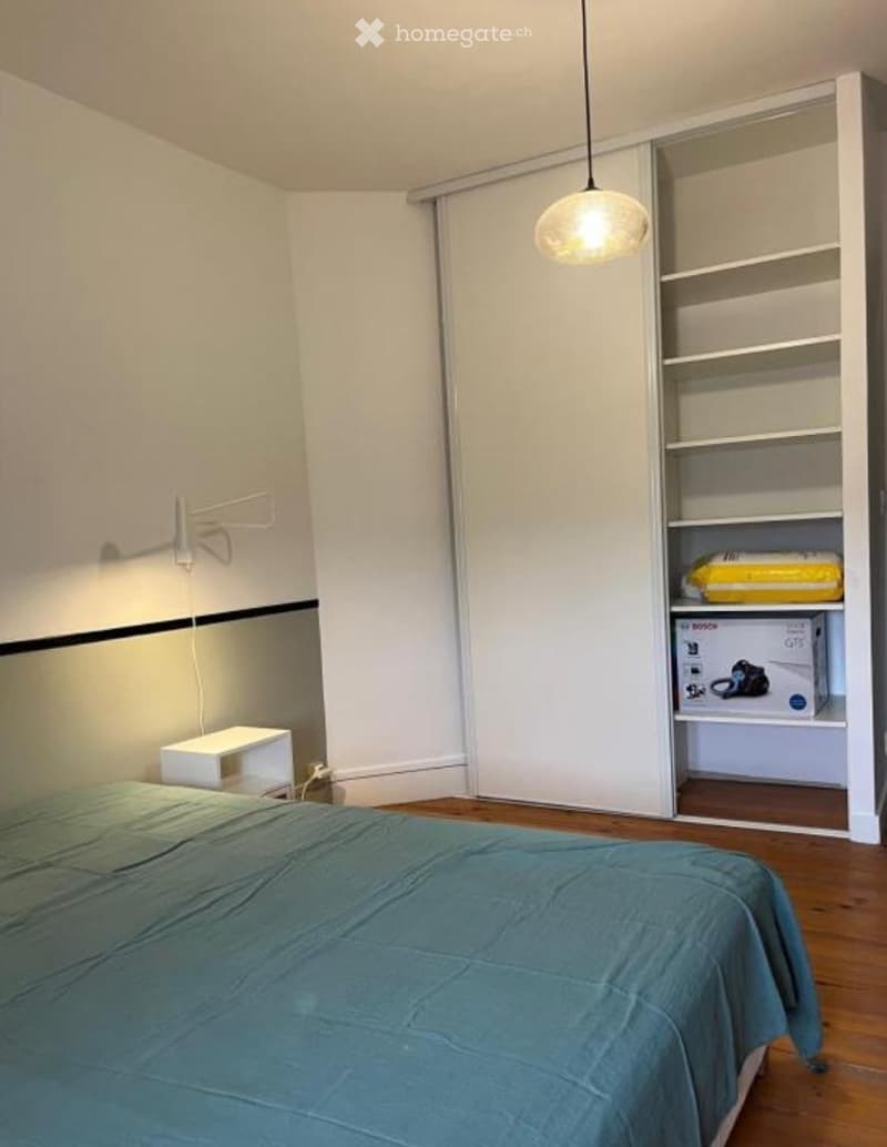 A modern bedroom with a neatly made bed, a nightstand, a built-in wardrobe with shelves, and a hanging pendant light.