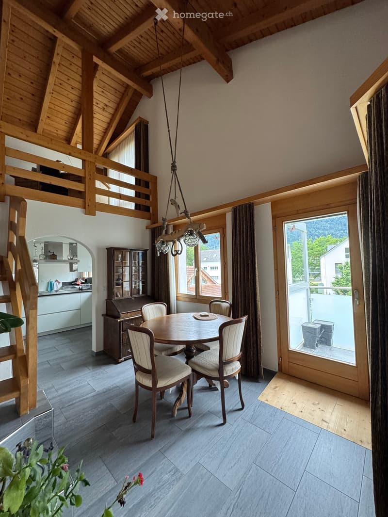 Dining area with a round wooden table and four chairs, a hanging chandelier, a kitchen area with cabinets and a sink, and large windows with curtains offering a view of the outdoors.