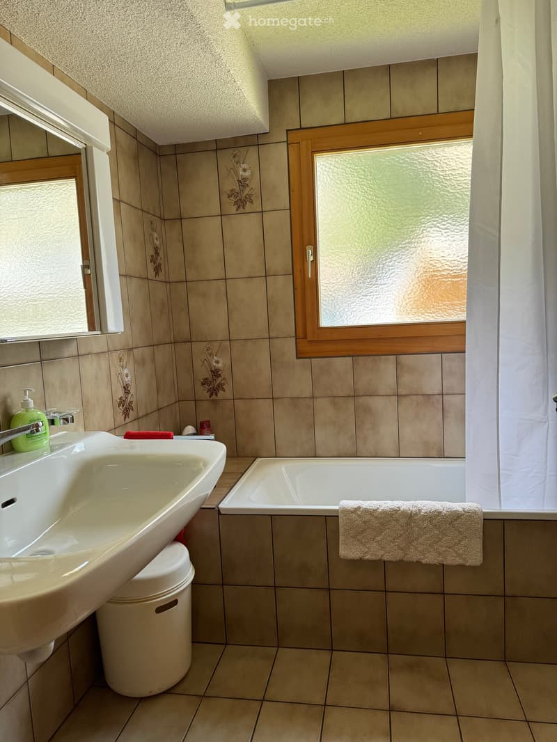 Bathroom with beige tiled walls and floor, a white sink with a chrome faucet, a bathtub, a towel on the edge, and a soap dispenser on the sink.