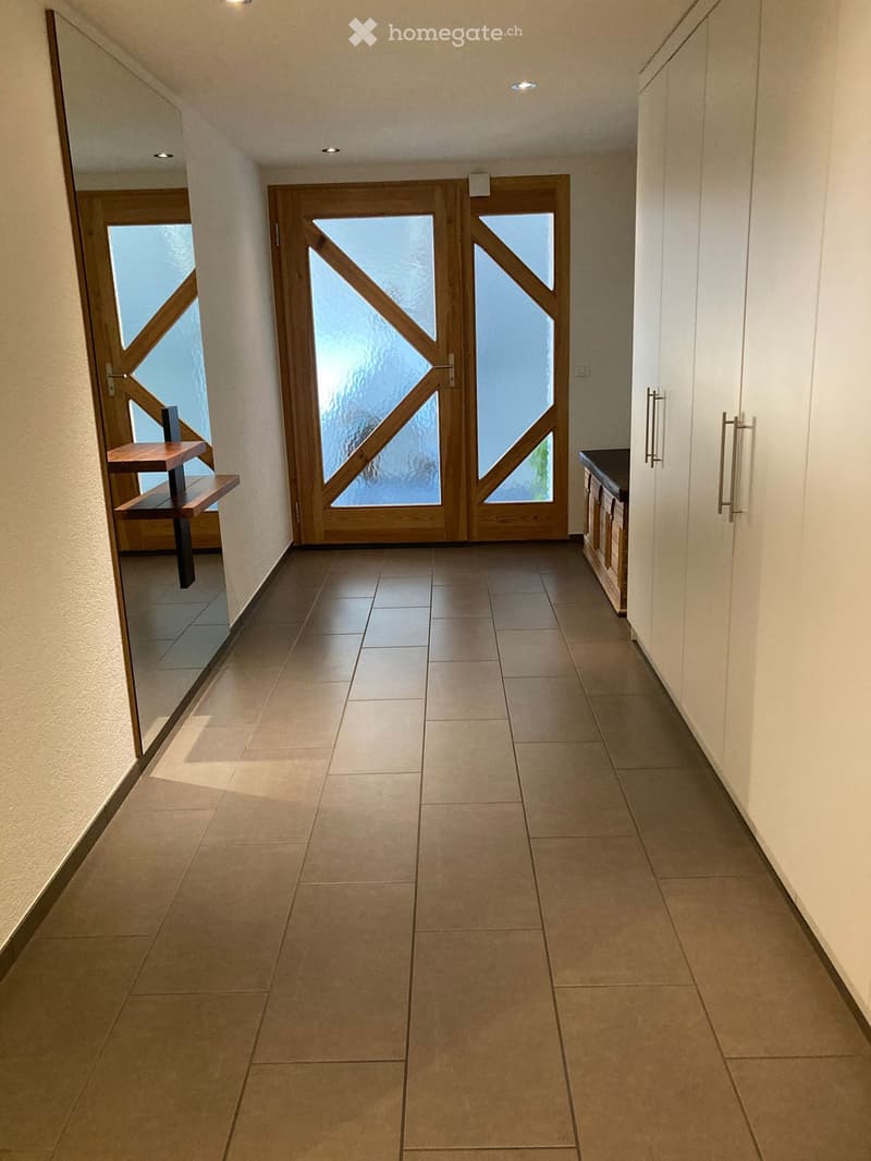 Modern hallway with tiled floor, recessed lighting, large wooden double doors with glass panels, and a small wooden bench to the left.