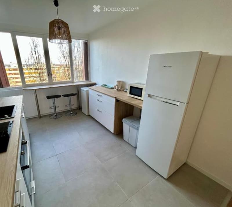 Modern kitchen with white cabinets, a refrigerator, microwave, and a kitchen island with stools. Large windows provide natural light.