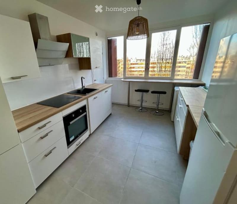 Modern kitchen with white cabinets, wooden countertops, built-in oven, sink, and stools at a small dining table. Large windows provide natural light.