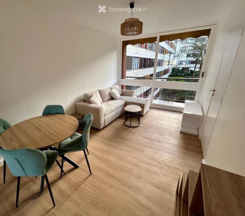 Bright living room with large windows and balcony access, featuring a light-colored sofa, coffee table, dining table with chairs, and a hanging floor lamp.