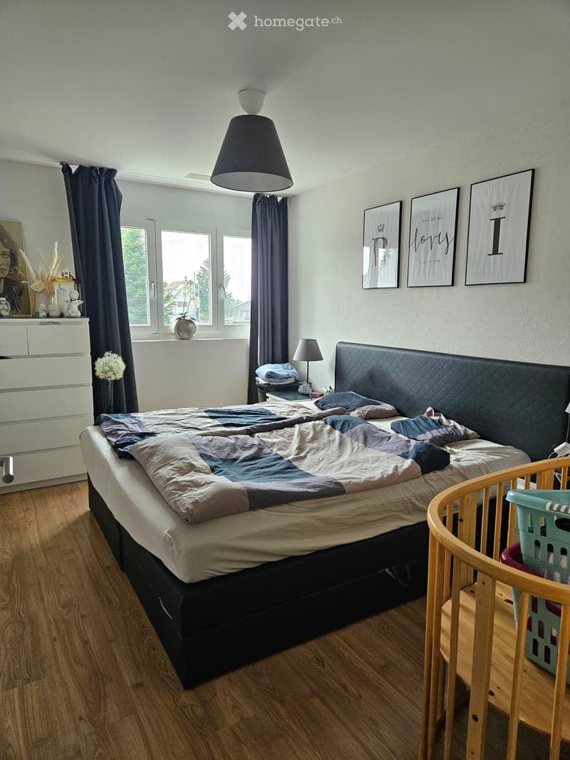 A well-lit bedroom with a large bed, nightstand, and a baby crib in the corner. The room features wooden flooring, large windows with dark curtains, and several decorative items including picture frames and a vase on the dresser.