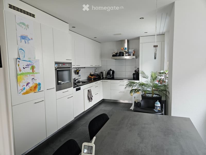 Modern kitchen with white cabinets, stainless steel appliances, and a grey countertop. Features include a refrigerator with children's drawings, an oven, a dishwasher, a coffee maker, a potted plant, and a dining area with a table and chairs.