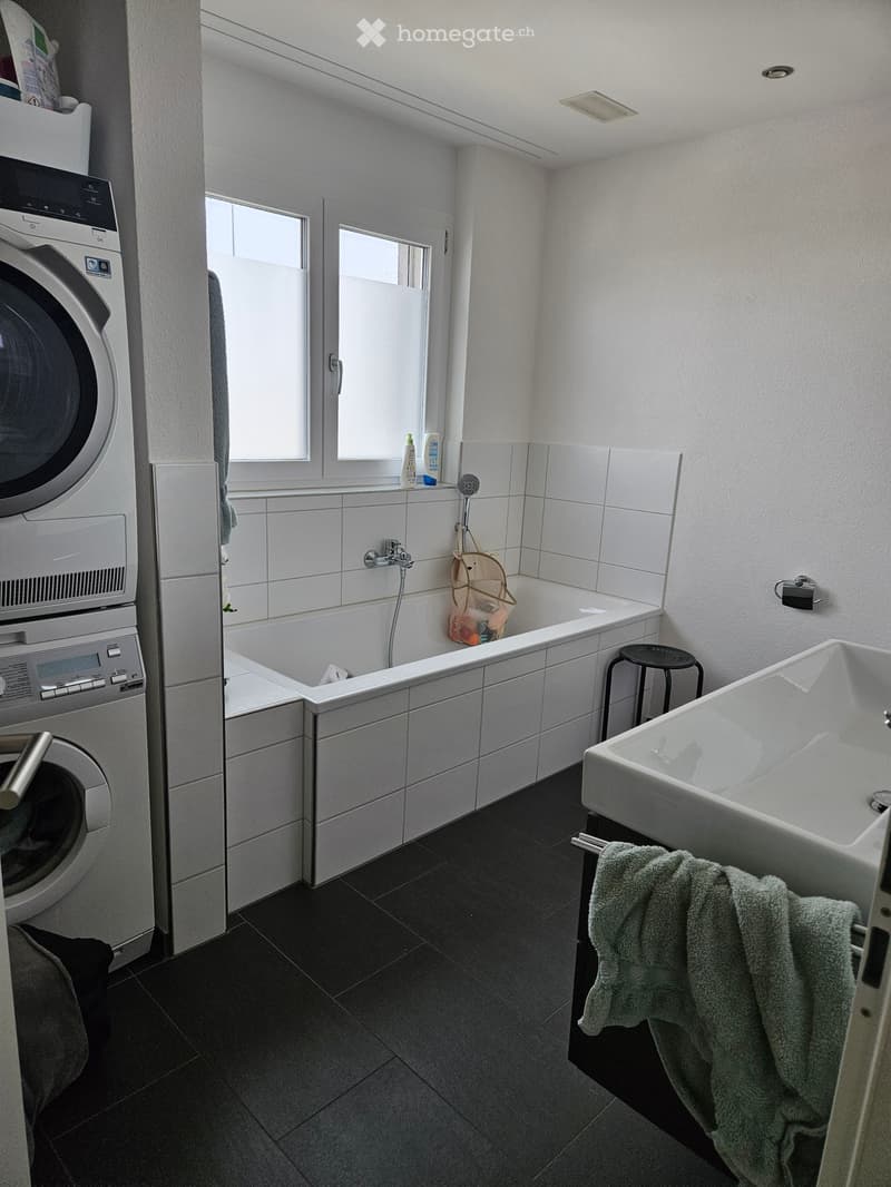 Modern bathroom with a bathtub, sink, washing machine, and a small stool. The space features dark tiled flooring and white tiled walls.