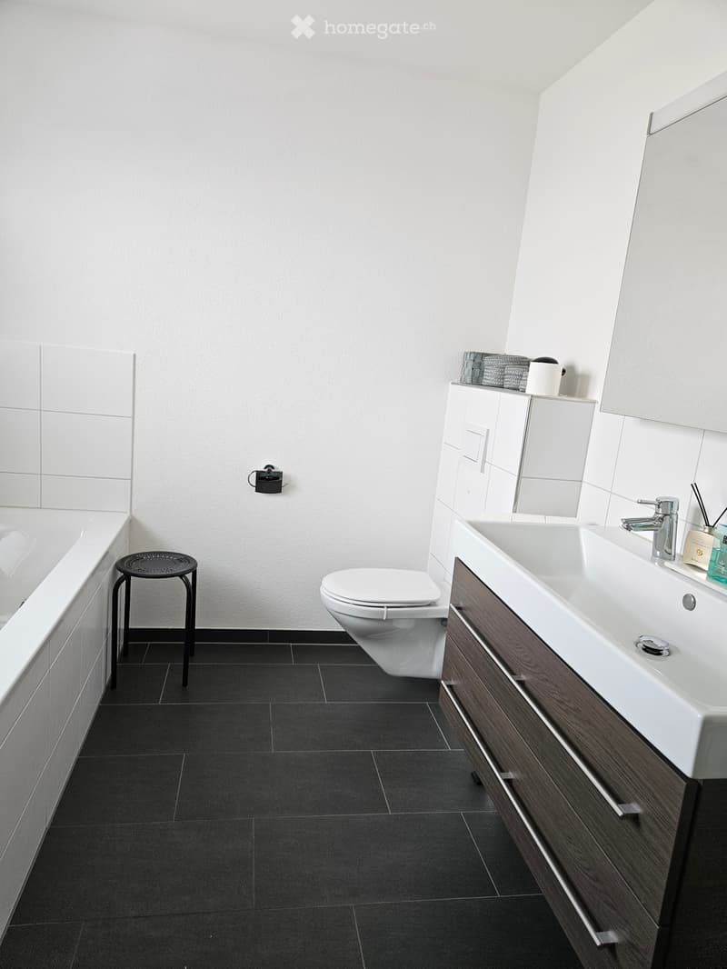 Modern bathroom with white tiles, dark floor tiles, a bathtub, toilet, sink, and a small black stool next to the bathtub.