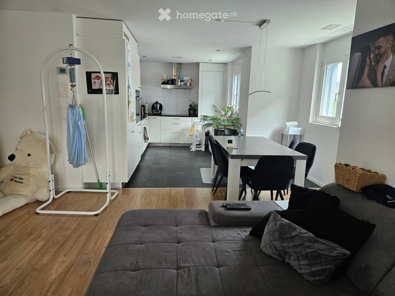 Open-plan living and dining area with a grey sofa, coffee table, dining table with chairs, kitchen area with cabinets and appliances, and a large teddy bear in the corner.