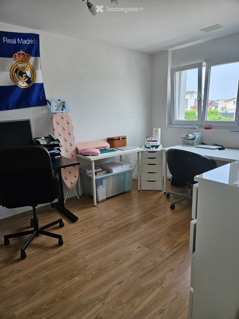 Office space with wooden flooring, two desks, chairs, a laptop, an ironing board, and storage bins.