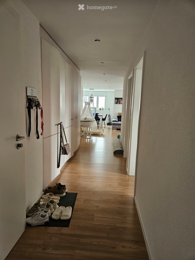 A narrow hallway with wooden flooring, leading to a room with a baby crib, dining table, and chairs. Shoes and bags are placed near the entrance.