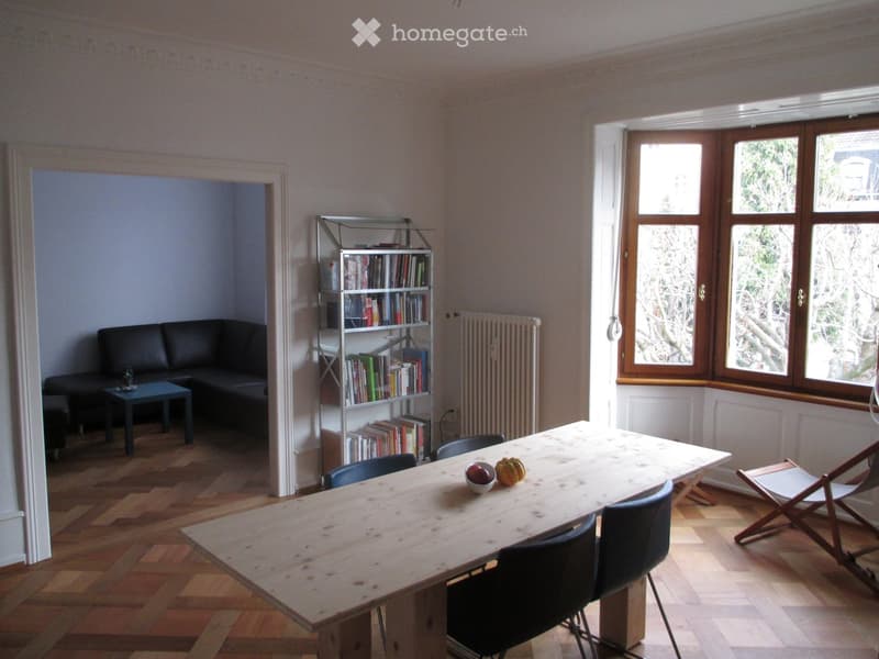 A modern living room with a wooden floor, large windows, a black leather sofa, a wooden dining table with chairs, and a bookshelf filled with books.