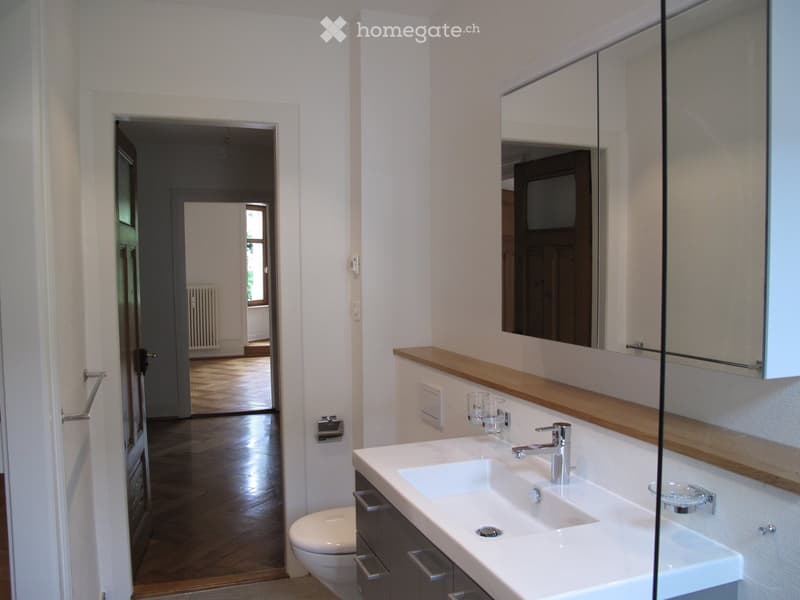Modern bathroom with a double sink, large mirror, and a toilet. The room features light-colored walls and wooden flooring.