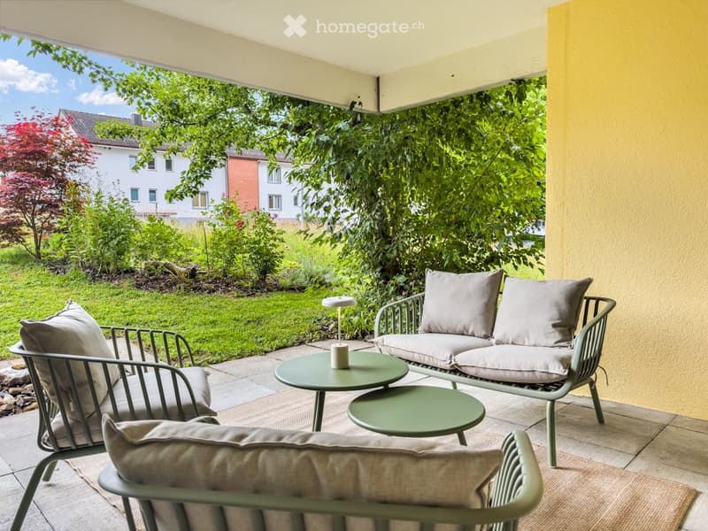 A modern balcony with a sofa, two chairs, two small tables, and a lamp, overlooking a well-maintained garden.
