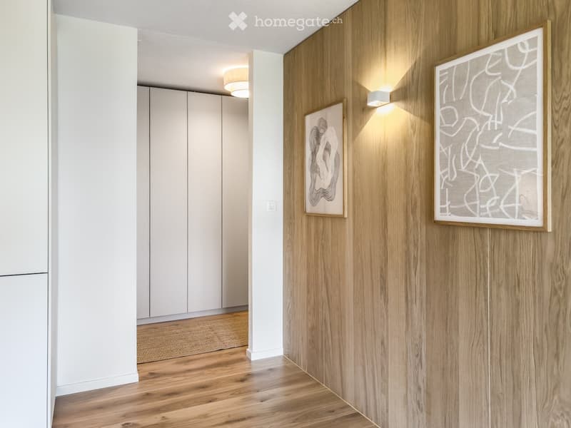 Modern hallway with wooden flooring, white cabinetry, and two abstract art pieces on the wall. Recessed lighting and a small rug are visible.