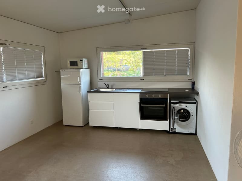 Compact kitchen with white cabinets, a sink, oven, refrigerator, microwave, and washing machine. Large window with blinds offers a view of greenery outside.
