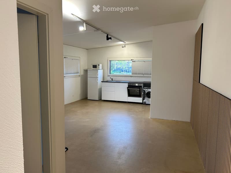 A small, unfurnished kitchen area with white cabinets, a refrigerator, microwave, oven, and washing machine. The space has a concrete floor and is connected to a hallway.