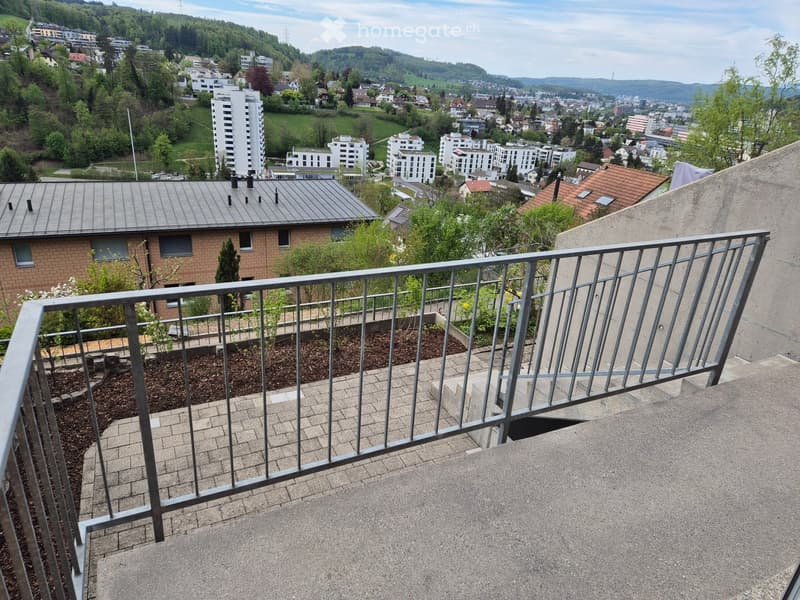 Balcony with metal railing overlooking a residential area with buildings, greenery, and distant hills.