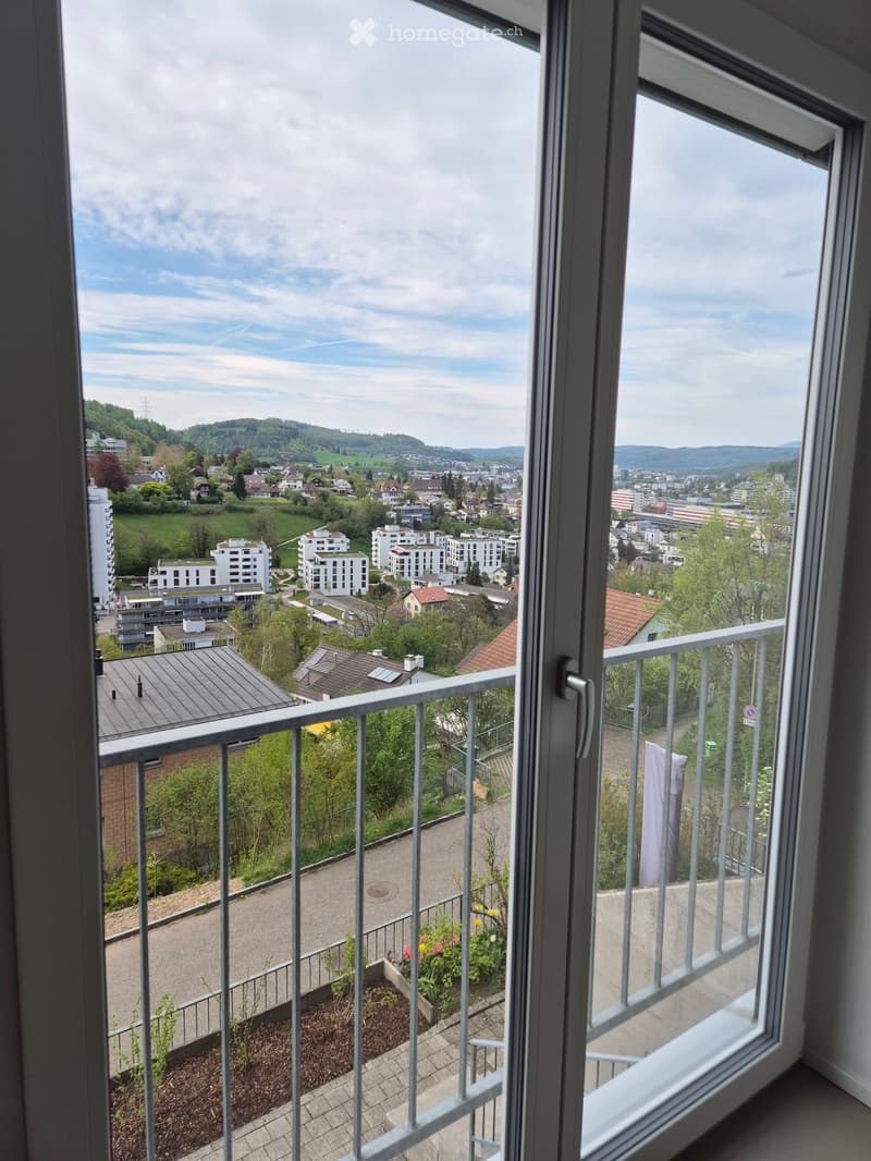 Balcony with metal railing, overlooking a cityscape with buildings, greenery, and distant hills under a partly cloudy sky.