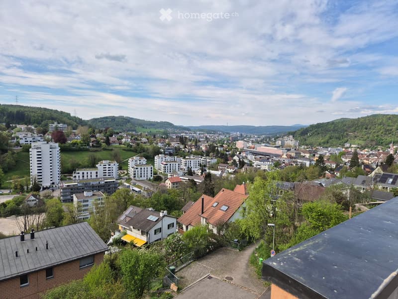 A panoramic view of a suburban area with a mix of residential buildings, including apartment complexes and single-family homes, surrounded by greenery and hills in the background.