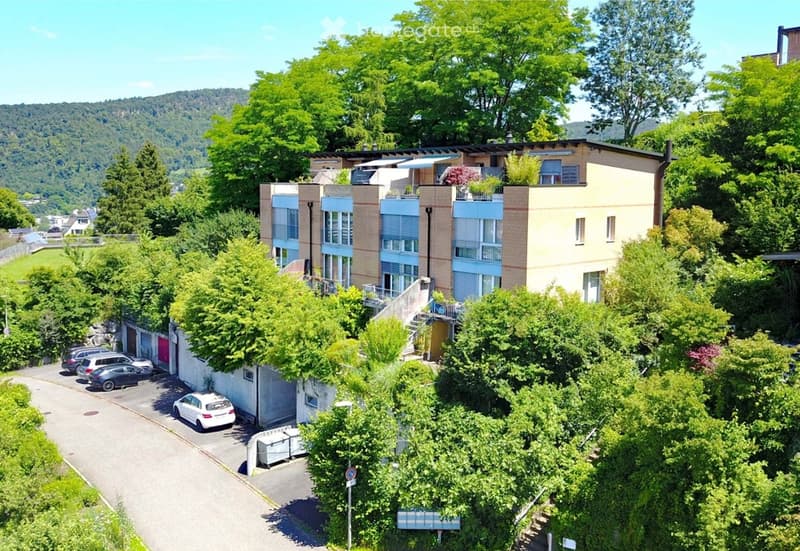 A modern multi-story residential building with balconies, surrounded by lush greenery and trees. Several cars are parked in the driveway.