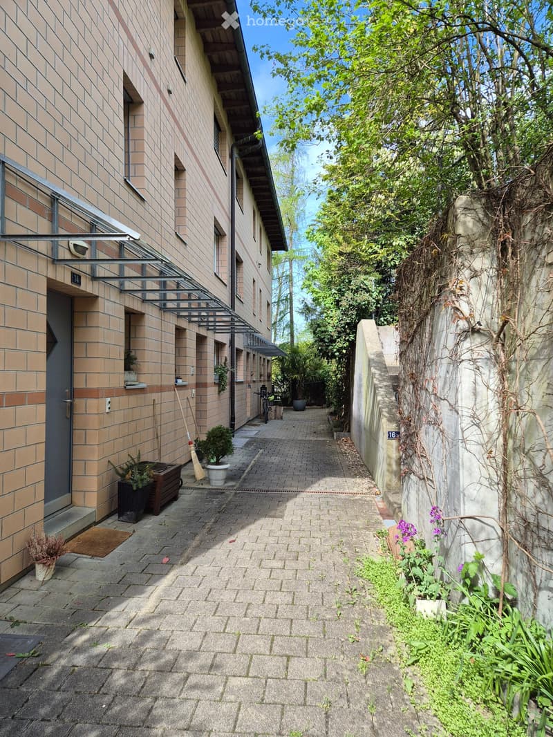 Narrow brick-paved alleyway between two buildings, with potted plants, bicycles, and greenery on both sides.