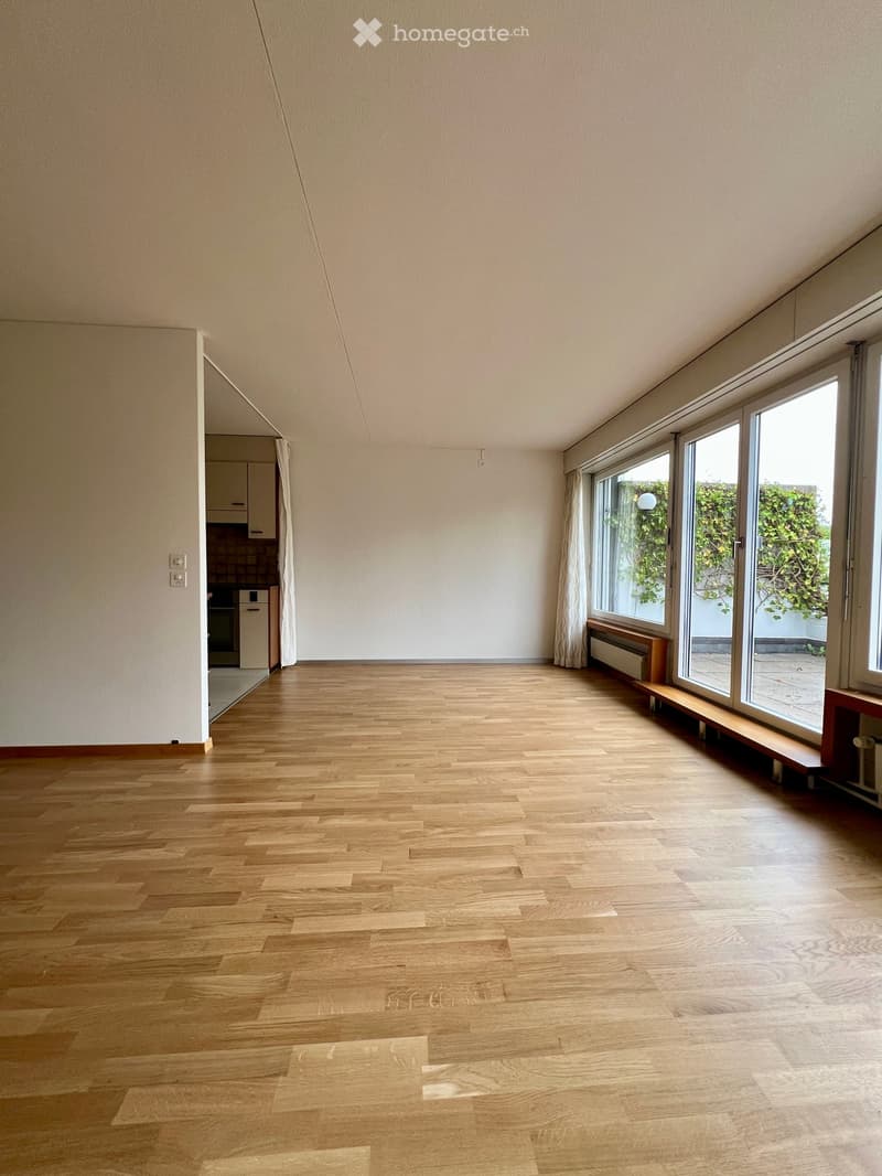 Empty room with wooden flooring, large windows, and a small bench near the window. The room has white walls and a visible kitchen area in the background.