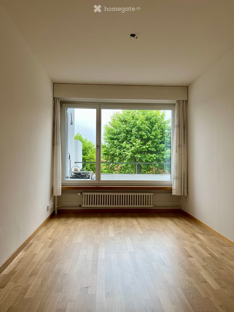 Empty room with light wooden flooring, white walls, large window with curtains, radiator, and a small balcony visible through the window.