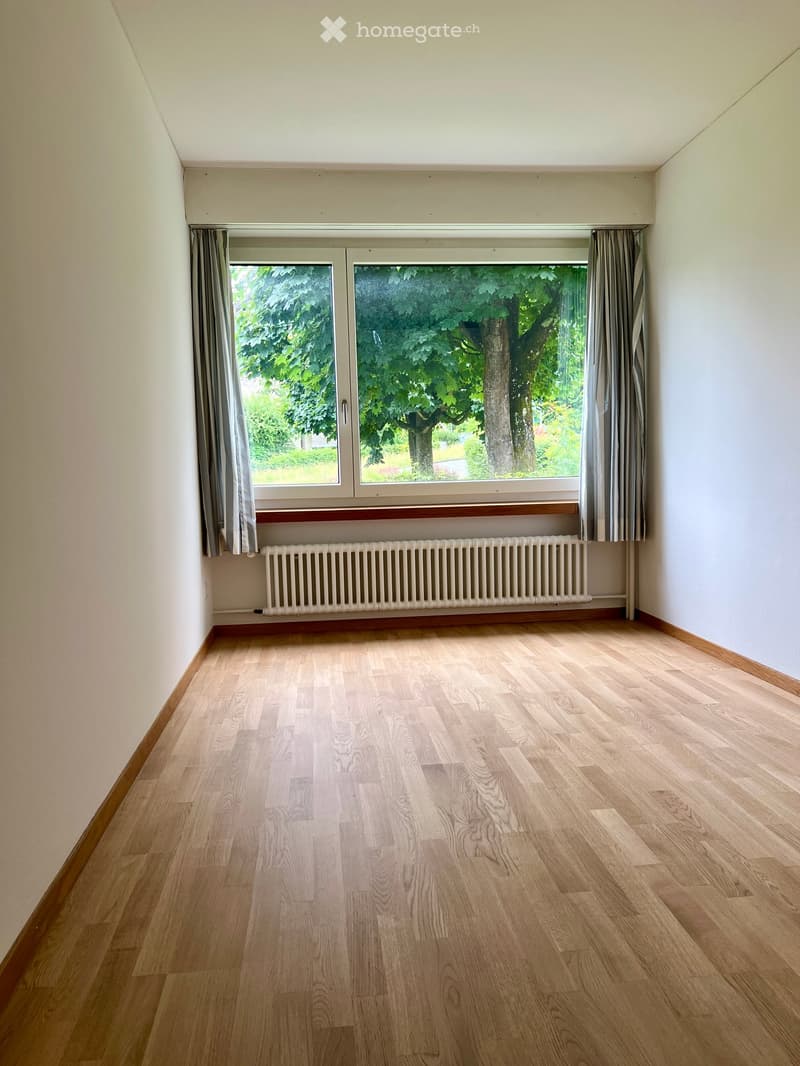 Empty room with light wooden flooring, white walls, large window with curtains, and a radiator below the window.