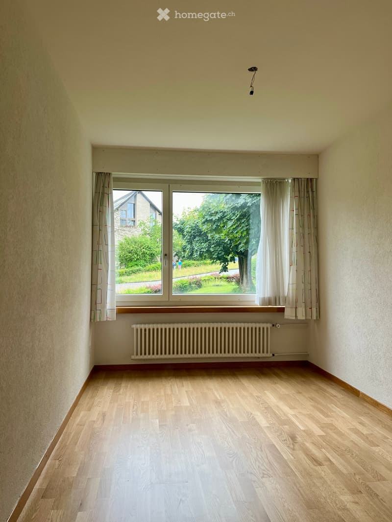 Empty room with wooden flooring, light-colored walls, large window with curtains, radiator below the window, and a view of greenery outside.