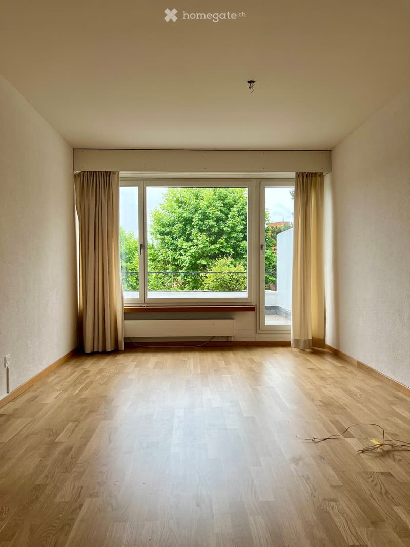 Empty room with wooden flooring, large windows with curtains, and a radiator below the windows.