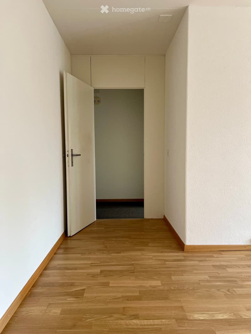Empty hallway with light wooden flooring, white walls, and a partially open door leading to another room.