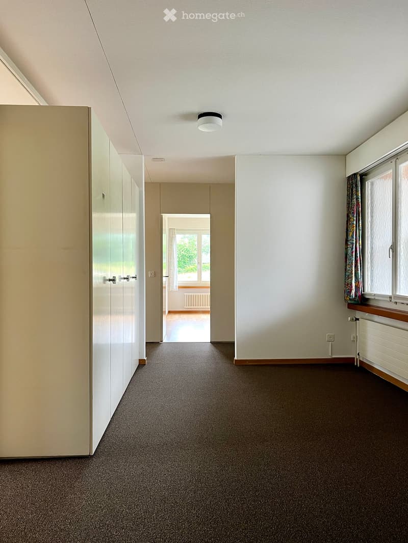 Empty hallway with carpeted floor, white walls, built-in closet, and a window with curtains. A doorway leads to another room with wooden flooring and large windows.
