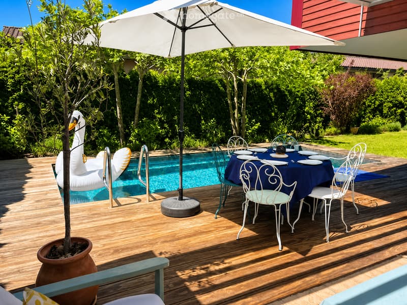 Outdoor patio area with a swimming pool, a dining table with chairs, and a large umbrella providing shade. There is a decorative swan float in the pool and a potted plant nearby.