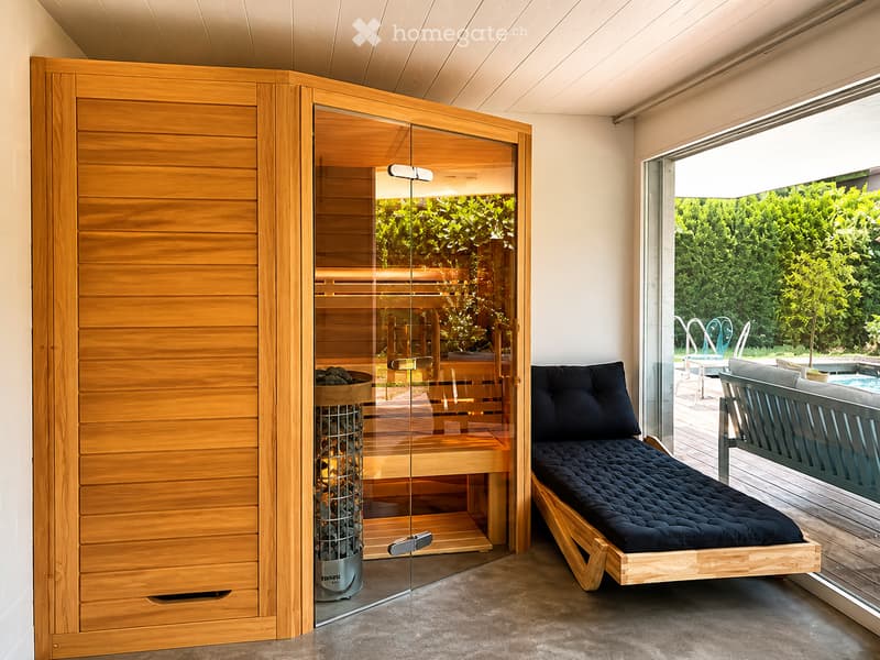 Modern indoor sauna room with a wooden sauna, a wooden bed, and a wooden cabinet. Large windows offer a view of the garden and outdoor pool area.