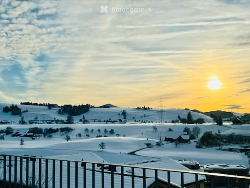 View from a balcony or terrace overlooking a snowy landscape with scattered houses and a sunset sky.
