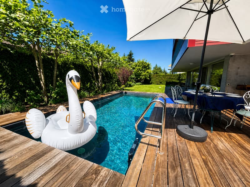 Outdoor area with a swimming pool, wooden decking, patio furniture, and a large umbrella. A swan-shaped inflatable float is in the pool.