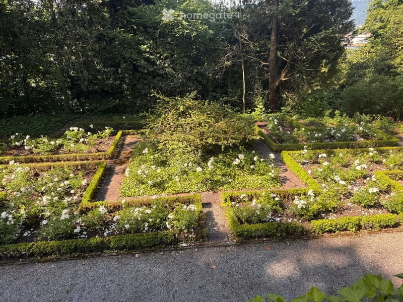 Formal garden with neatly trimmed hedges, flower beds, and gravel pathways surrounded by trees and greenery.