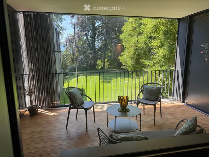Modern balcony with wooden decking, two chairs, a small table, a sofa, and a decorative vase. Large windows offer a view of a green lawn and trees.