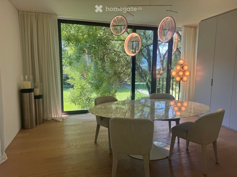 Modern dining room with a round glass table, four upholstered chairs, large windows with curtains, and decorative lighting fixtures.