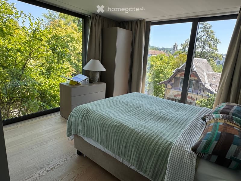 Modern bedroom with large windows offering a scenic view, featuring a neatly made bed, nightstand with a lamp, and a book.