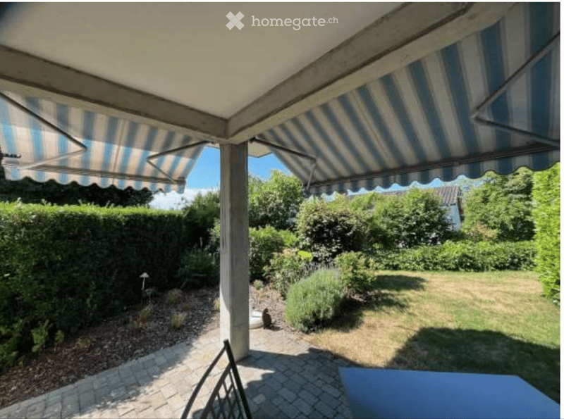 A covered outdoor patio area with striped awnings, leading to a well-maintained garden with a lawn, shrubs, and a gravel pathway.
