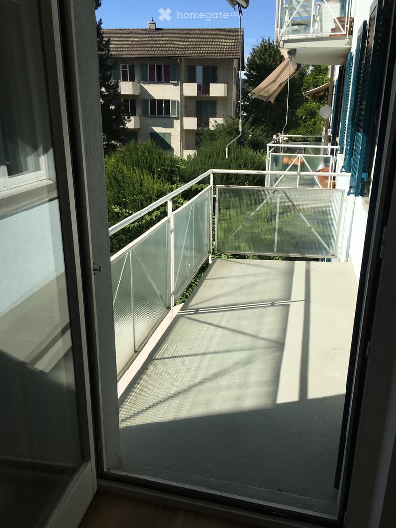 Balcony with glass railing, overlooking a residential area with trees and neighboring buildings.